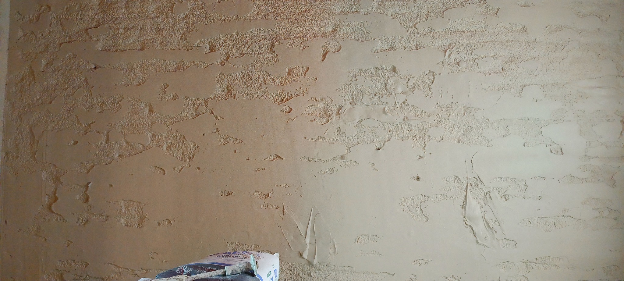 Teksturowana, beżowa ściana po nałożeniu tynku dekoracyjnego. Widoczne nierówności i wzory. W dolnej części worki z zaprawą i narzędzia.