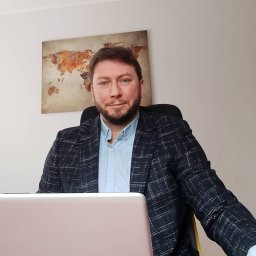 Łukasz Kozłowski - Doradca kredytowy w marynarce w kratę siedzi za biurkiem z laptopem, w tle mapa świata. Uśmiechnięty, kontaktowy, budzący zaufanie.