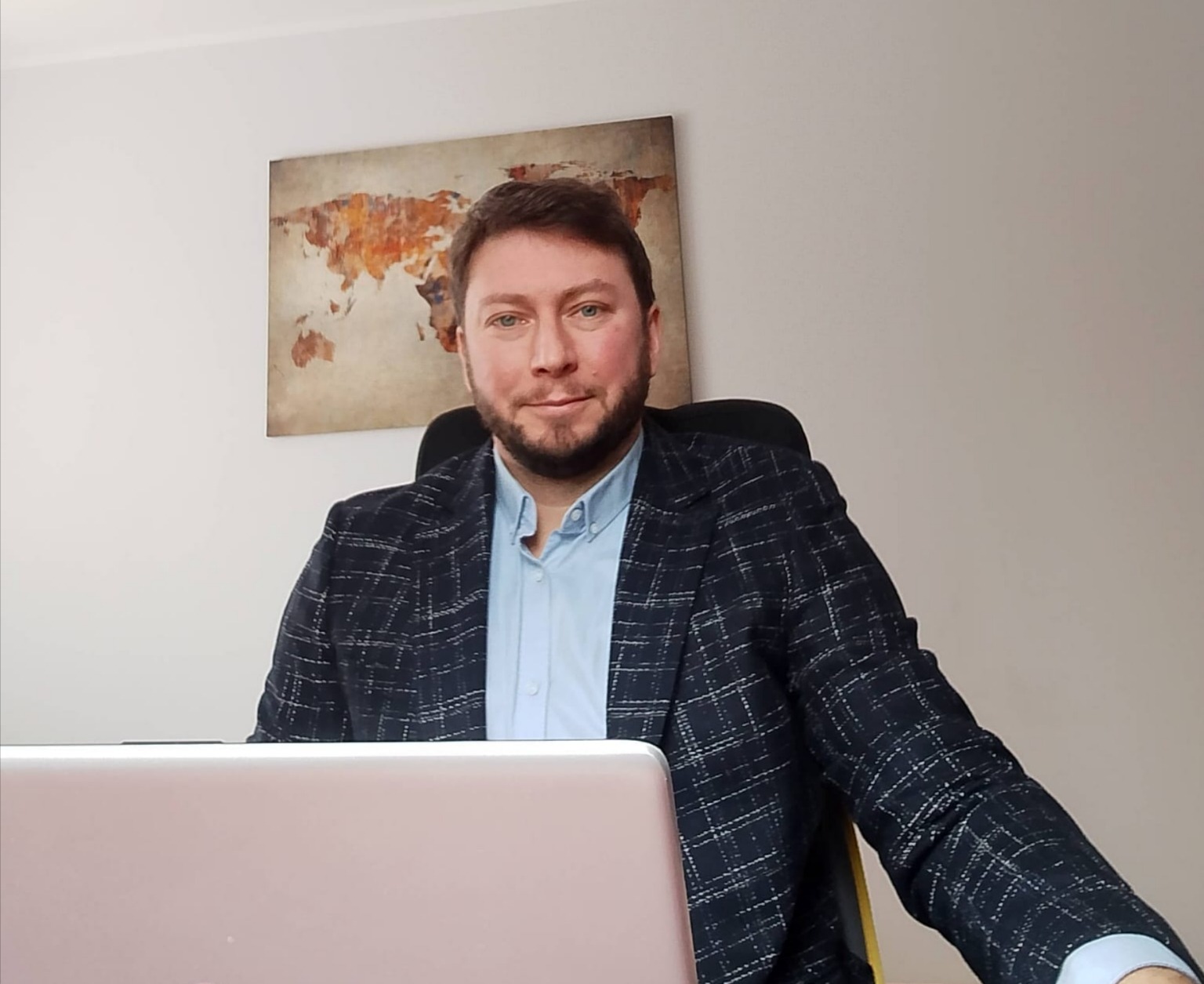 Doradca kredytowy w marynarce w kratę siedzi za biurkiem z laptopem, w tle mapa świata. Uśmiechnięty, kontaktowy, budzący zaufanie.