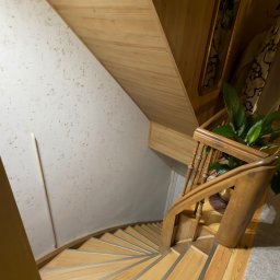 Krzysztof Chmiel Serwis Domowy Naprawy i Remonty - Drewniane schody spiralne z jasnego drewna, widok z góry. Ściana z dekoracyjną fakturą, lustro w drewnianej ramie i roślina doniczkowa dodają wnętrzu przytulności.