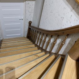 Krzysztof Chmiel Serwis Domowy Naprawy i Remonty - Drewniane schody z metalowymi listwami antypoślizgowymi, białe drzwi w tle, ściana z fakturą. Ujęcie z góry, widoczne detale wykończenia.