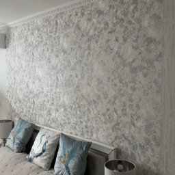 Krzysztof Chmiel Serwis Domowy Naprawy i Remonty - Ściana w sypialni pokryta dekoracyjną, srebrzysto-białą tapetą w kwiatowe wzory. Widoczny fragment łóżka z poduszkami i obraz na ścianie. Górna krawędź ściany wykończona sztukaterią.