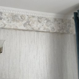 Krzysztof Chmiel Serwis Domowy Naprawy i Remonty - Eleganckie wnętrze: ściana z tapetą w odcieniach szarości, ozdobny pas z motywem kwiatowym pod sufitem, kinkiet i zasłony w kolorze morskim.