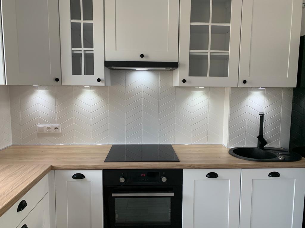 Biała kuchnia z drewnianym blatem i płytkami ułożonymi w jodełkę. Nowoczesny design z czarnymi akcentami, oświetlenie punktowe pod szafkami.