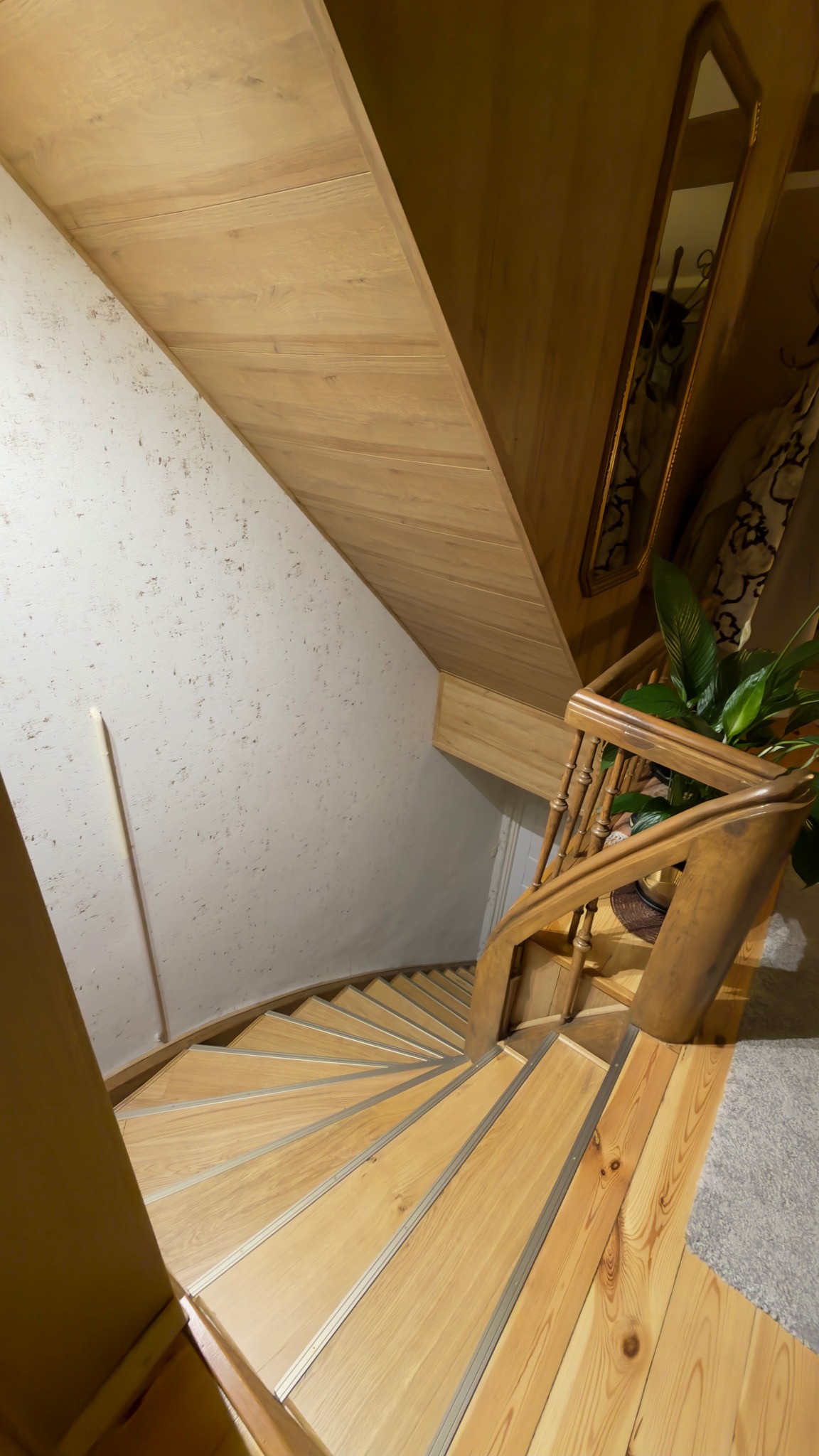 Drewniane schody spiralne z jasnego drewna, widok z góry. Ściana z dekoracyjną fakturą, lustro w drewnianej ramie i roślina doniczkowa dodają wnętrzu przytulności.