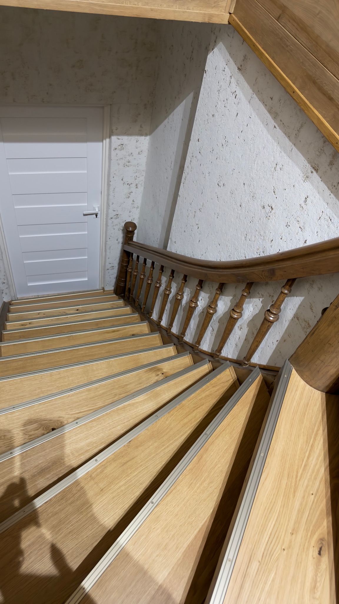 Drewniane schody z metalowymi listwami antypoślizgowymi, białe drzwi w tle, ściana z fakturą. Ujęcie z góry, widoczne detale wykończenia.