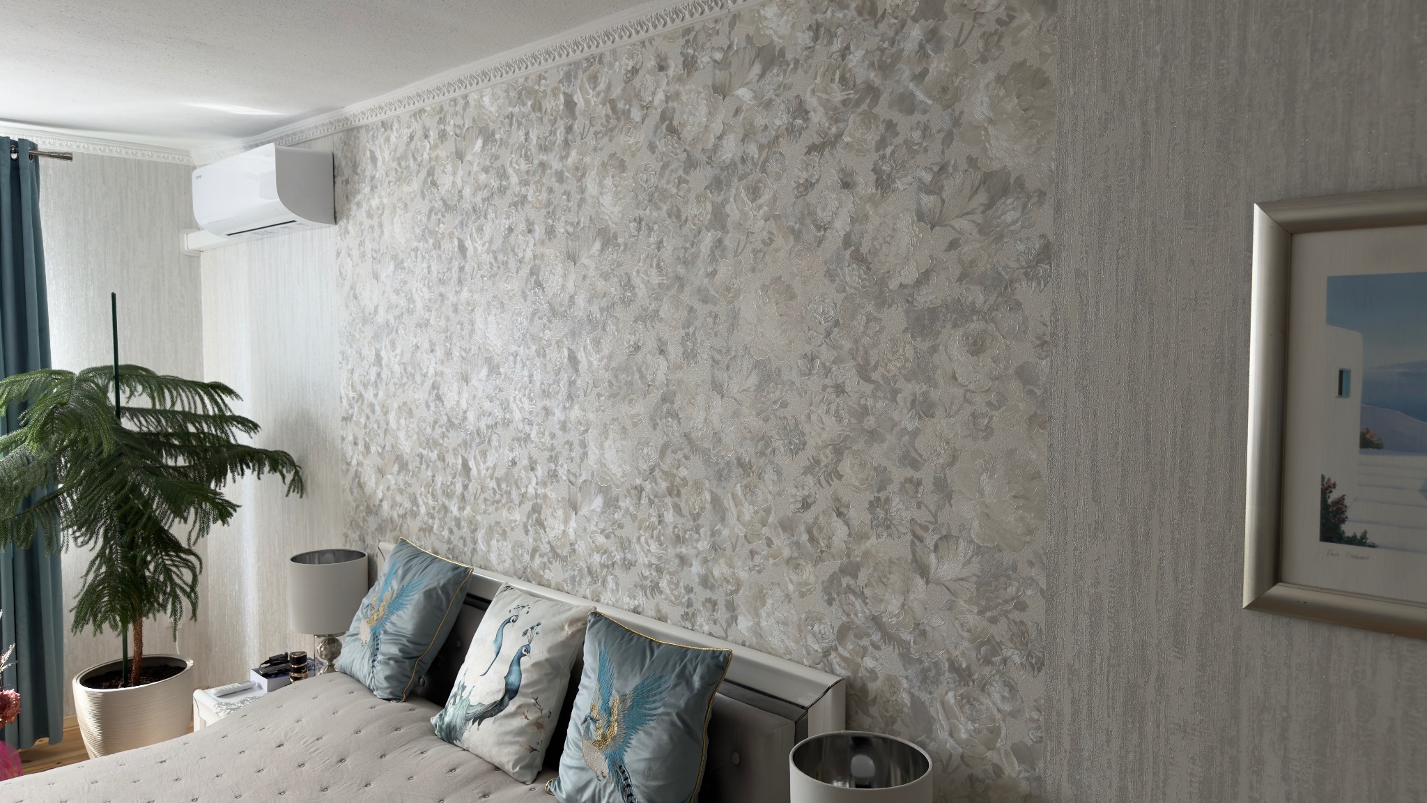 Ściana w sypialni pokryta dekoracyjną, srebrzysto-białą tapetą w kwiatowe wzory. Widoczny fragment łóżka z poduszkami i obraz na ścianie. Górna krawędź ściany wykończona sztukaterią.