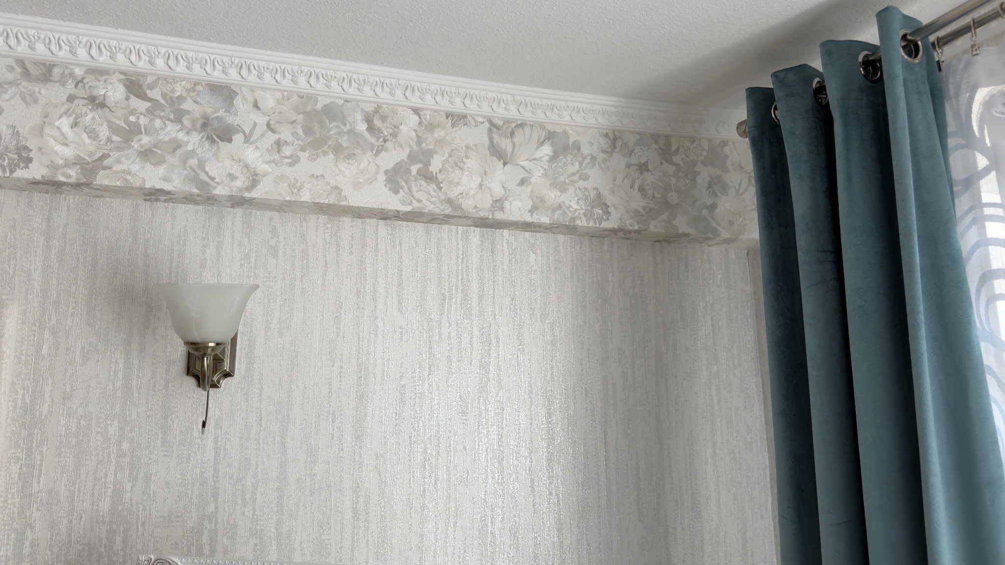 Eleganckie wnętrze: ściana z tapetą w odcieniach szarości, ozdobny pas z motywem kwiatowym pod sufitem, kinkiet i zasłony w kolorze morskim.