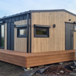MODERAHOUSE SPÓŁKA Z OGRANICZONĄ ODPOWIEDZIALNOŚCIĄ - Gotowy dom modułowy z drewnianą elewacją i tarasem, czarne okna i drzwi, klimatyzator na ścianie, widok z zewnątrz na tle zabudowań gospodarczych i drzew.