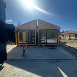 MODERAHOUSE SPÓŁKA Z OGRANICZONĄ ODPOWIEDZIALNOŚCIĄ - Dwa moduły domu z drewna z dużymi oknami, ustawione obok siebie na betonowej powierzchni, w tle budynek gospodarczy i błękitne niebo z mocnym słońcem.