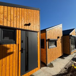 MODERAHOUSE SPÓŁKA Z OGRANICZONĄ ODPOWIEDZIALNOŚCIĄ - Nowoczesne domy modułowe z drewnianą elewacją w odcieniach pomarańczy i czarnymi oknami, ustawione na fundamencie, w słoneczny dzień.