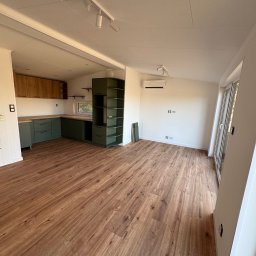 MODERAHOUSE SPÓŁKA Z OGRANICZONĄ ODPOWIEDZIALNOŚCIĄ - Wnętrze modułowego domu z drewnianą podłogą, zieloną zabudową kuchenną i oświetleniem punktowym. Widok na okno i klimatyzator na białej ścianie.