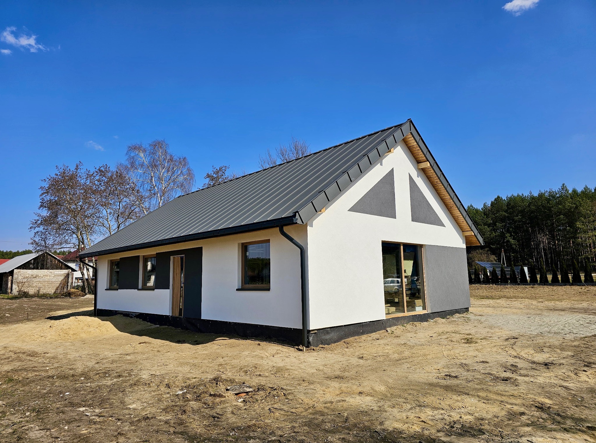 Nowoczesny dom modułowy z szarą blachodachówką i białymi ścianami, z elementami szarego tynku. Widoczne okna i drzwi. Dom otoczony piaszczystym terenem.
