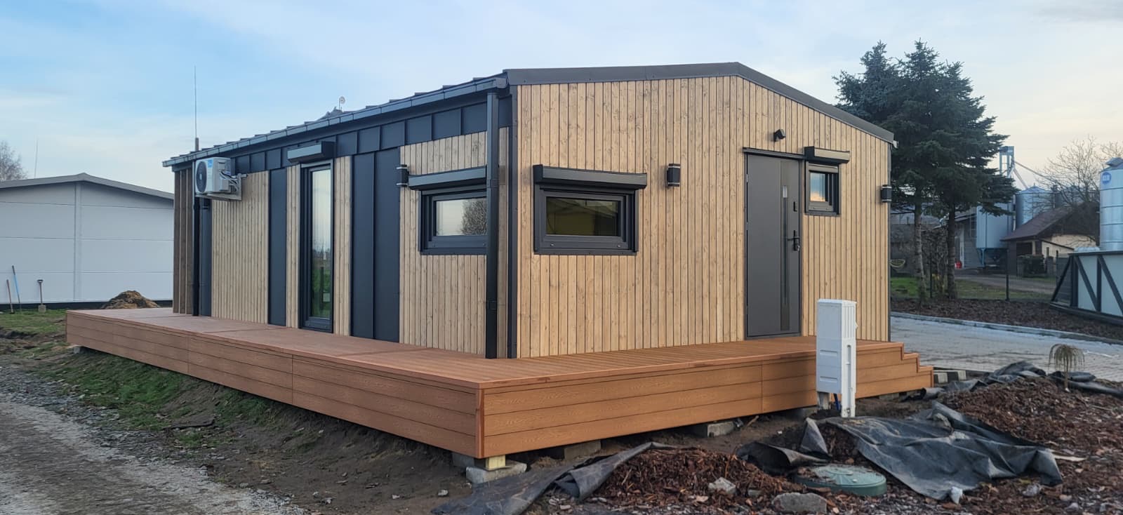 Gotowy dom modułowy z drewnianą elewacją i tarasem, czarne okna i drzwi, klimatyzator na ścianie, widok z zewnątrz na tle zabudowań gospodarczych i drzew.