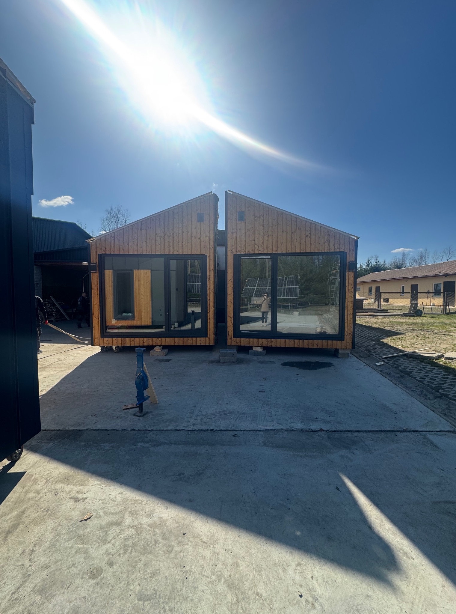 Dwa moduły domu z drewna z dużymi oknami, ustawione obok siebie na betonowej powierzchni, w tle budynek gospodarczy i błękitne niebo z mocnym słońcem.