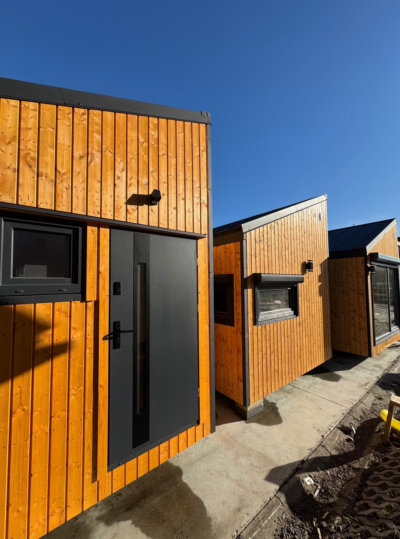 Nowoczesne domy modułowe z drewnianą elewacją w odcieniach pomarańczy i czarnymi oknami, ustawione na fundamencie, w słoneczny dzień.