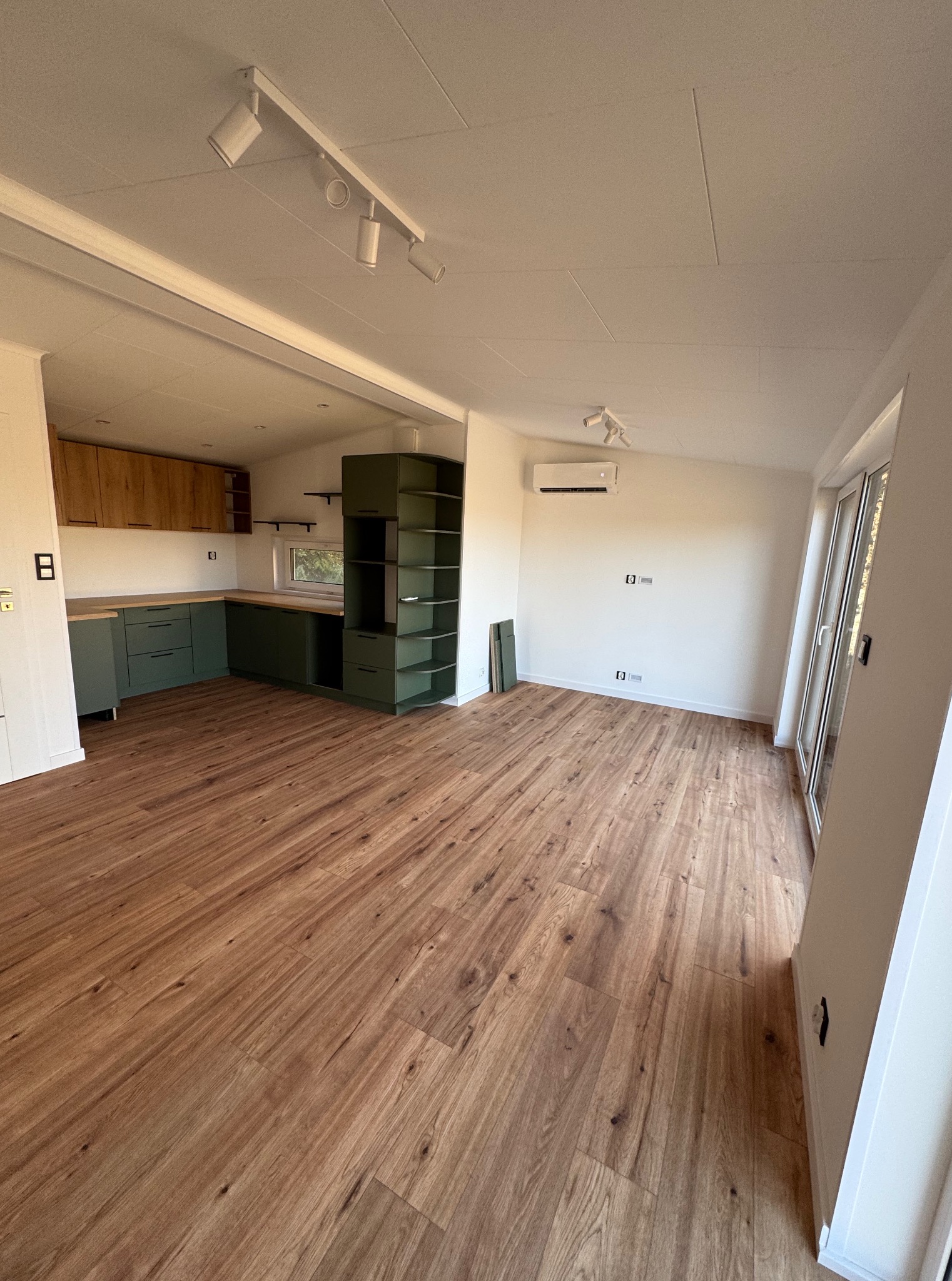 Wnętrze modułowego domu z drewnianą podłogą, zieloną zabudową kuchenną i oświetleniem punktowym. Widok na okno i klimatyzator na białej ścianie.