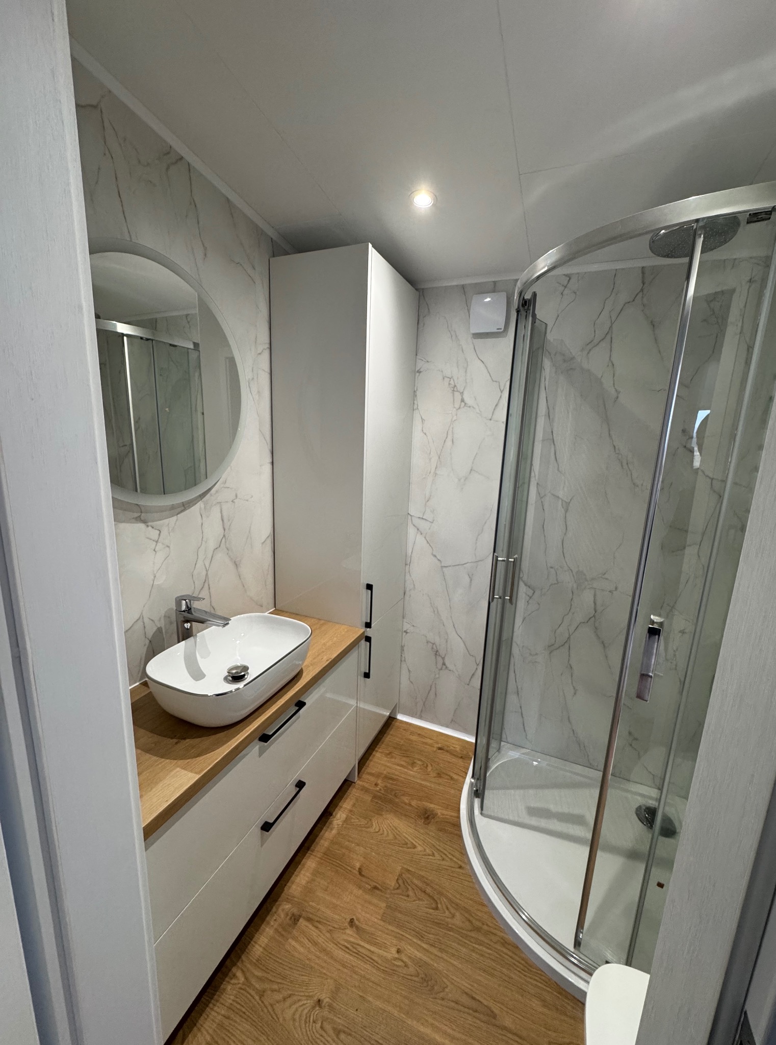 Nowoczesna łazienka w domu modułowym: kabina prysznicowa, umywalka nablatowa na szafce z drewnianym blatem, okrągłe lustro i marmurowe płytki ścienne.