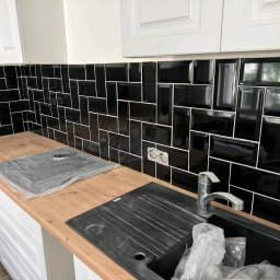 MG Wykonczenia Wnętrz - Czarne, błyszczące płytki ułożone w cegiełkę na ścianie nad blatem kuchennym. Widoczne gniazdka elektryczne i zlew. Wykończenie wnętrza kuchni w nowoczesnym stylu.