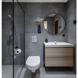 MG Wykonczenia Wnętrz - Nowoczesna łazienka w szarościach z kabiną prysznicową typu walk-in, okrągłym lustrem i drewnianą szafką pod umywalką. Minimalistyczny design z akcentem kwiatowym.