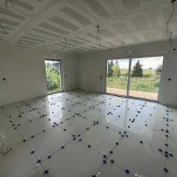 MG Wykonczenia Wnętrz - Wnętrze w trakcie wykańczania: białe płytki podłogowe z niebieskimi krzyżykami dystansowymi, sufit z płyt karton-gips, okna z widokiem na zieleń. Przewody elektryczne zwisają z sufitu.