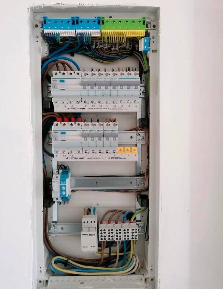 Rozdzielnica elektryczna Hager w białej obudowie, z precyzyjnie ułożonymi przewodami i zabezpieczeniami. Widoczne listwy zaciskowe i oznaczenia faz. Estetyczny montaż.