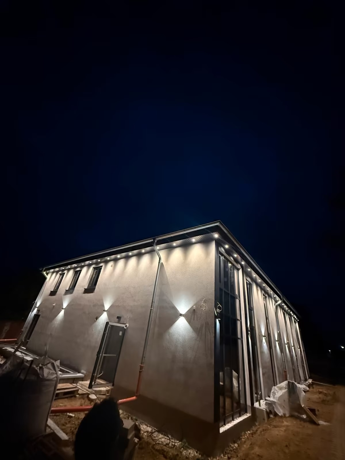 Oświetlona elewacja budynku nocą, z widocznymi lampami elewacyjnymi i listwami LED pod dachem. Budynek w trakcie wykończenia, widoczne materiały budowlane.