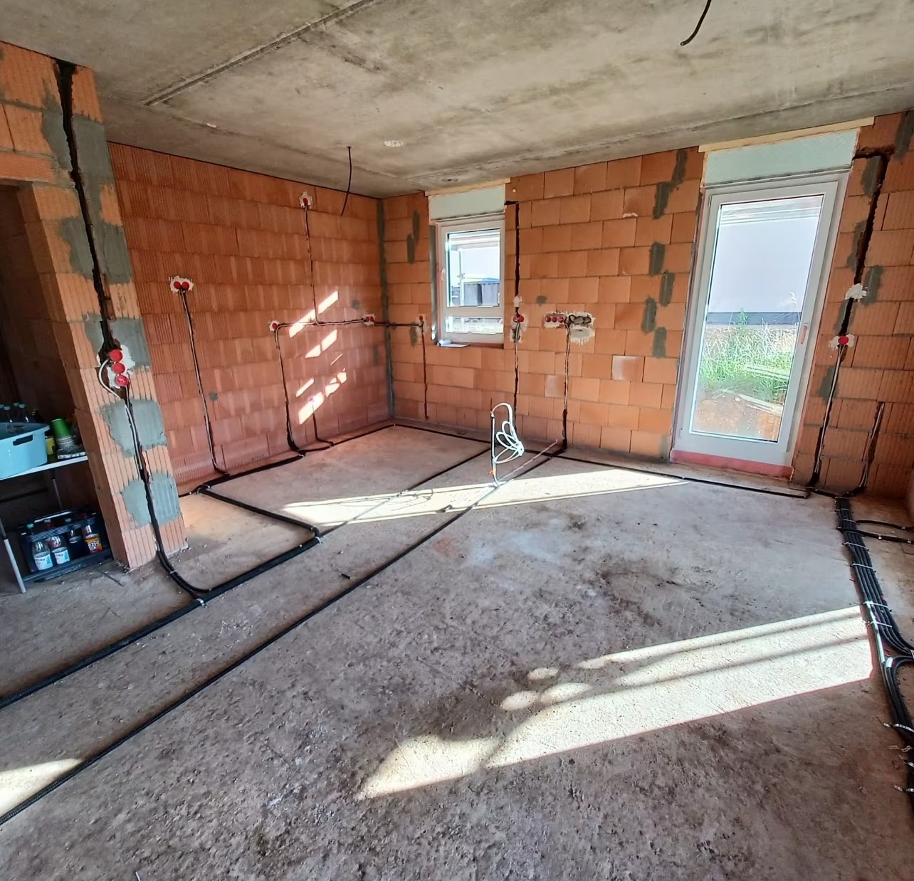 Surowy pokój z ceglanymi ścianami w trakcie instalacji elektrycznej: widoczne kable w bruzdach i puszki. Okno i drzwi balkonowe w tle. Podłoga betonowa.