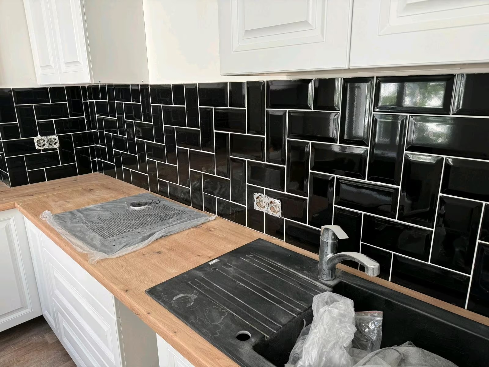 Czarne, błyszczące płytki ułożone w cegiełkę na ścianie nad blatem kuchennym. Widoczne gniazdka elektryczne i zlew. Wykończenie wnętrza kuchni w nowoczesnym stylu.