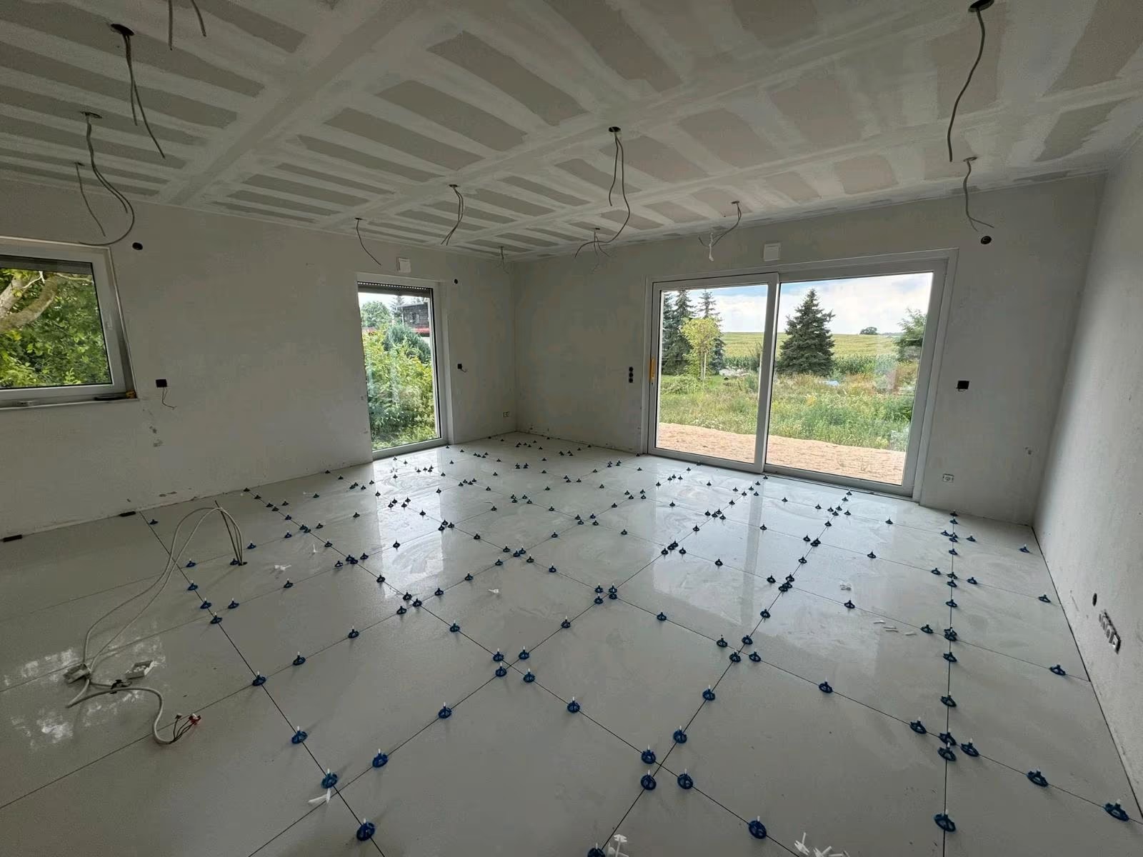 Wnętrze w trakcie wykańczania: białe płytki podłogowe z niebieskimi krzyżykami dystansowymi, sufit z płyt karton-gips, okna z widokiem na zieleń. Przewody elektryczne zwisają z sufitu.