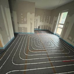 Krystian Binkowski - Układanie białych rur ogrzewania podłogowego na szarej macie w surowym wnętrzu z oknem. Widoczne ślady po gładzi na ścianach i wystające kable.