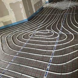 Krystian Binkowski - Białe rury ogrzewania podłogowego ułożone na folii z siatką, zabezpieczone taśmą. Widok z góry na instalację w surowym wnętrzu przed wylewką.