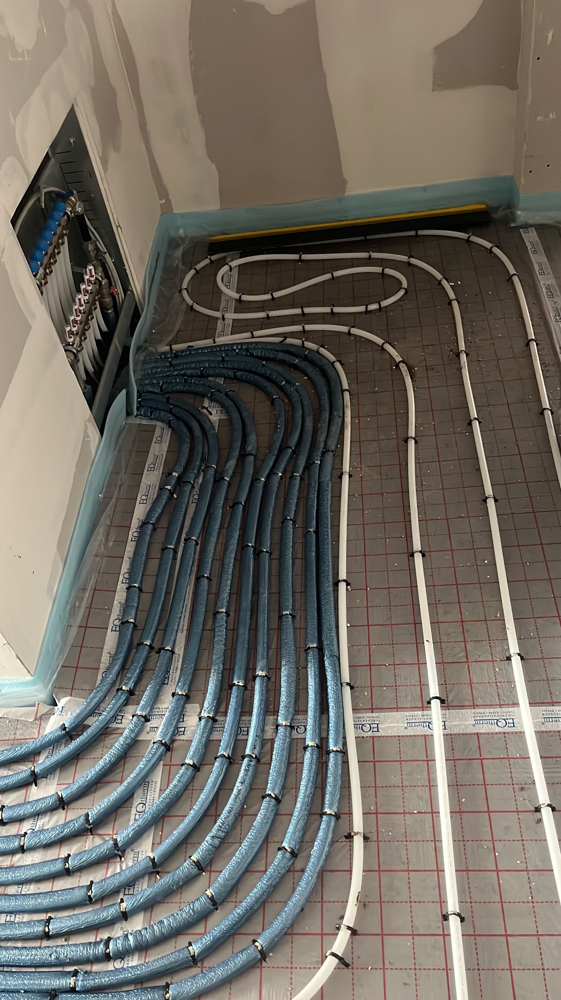 Niebieskie i białe rury ogrzewania podłogowego, przymocowane do podłoża z folią w kratkę, widoczne kolektory w ścianie. Instalacja w trakcie montażu.