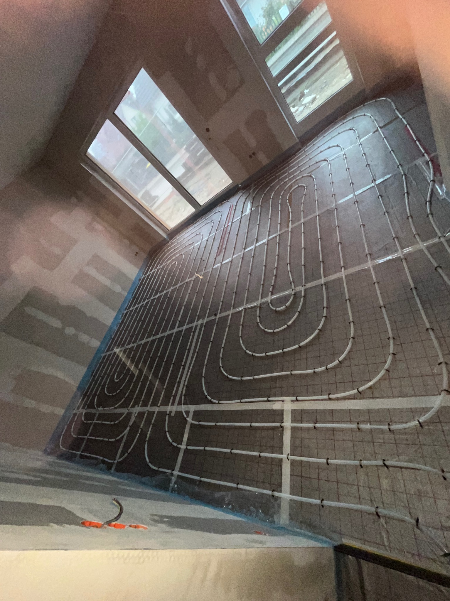 Białe rurki ogrzewania podłogowego ułożone na siatce w surowym wnętrzu z oknami dachowymi. Widoczne narzędzia i elementy instalacji.