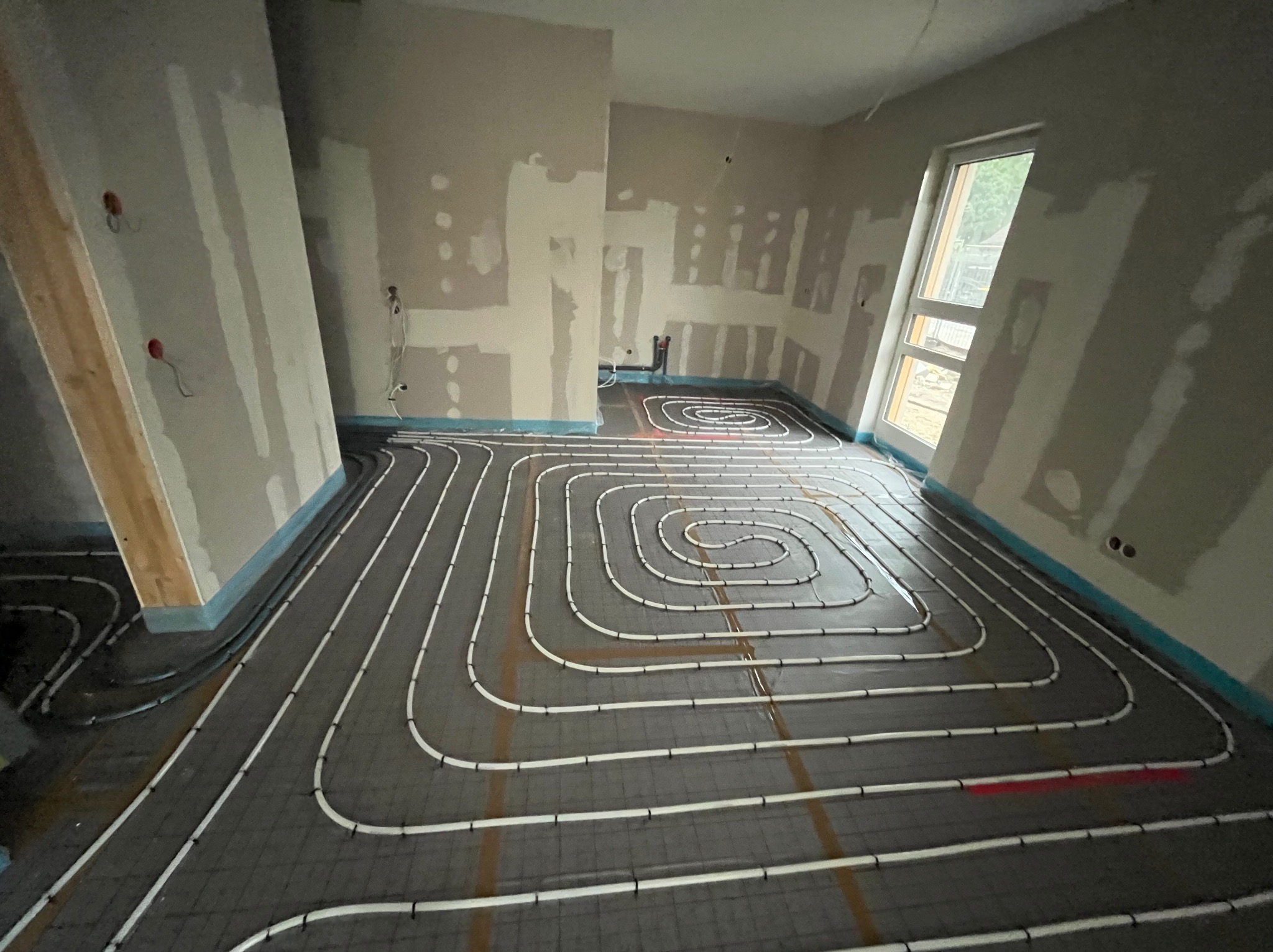 Układanie białych rur ogrzewania podłogowego na szarej macie w surowym wnętrzu z oknem. Widoczne ślady po gładzi na ścianach i wystające kable.