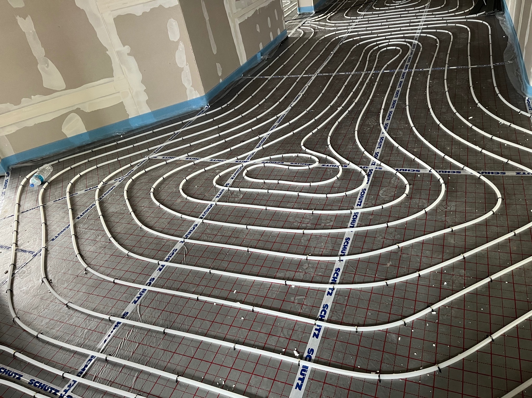 Białe rury ogrzewania podłogowego ułożone na folii z siatką, zabezpieczone taśmą. Widok z góry na instalację w surowym wnętrzu przed wylewką.