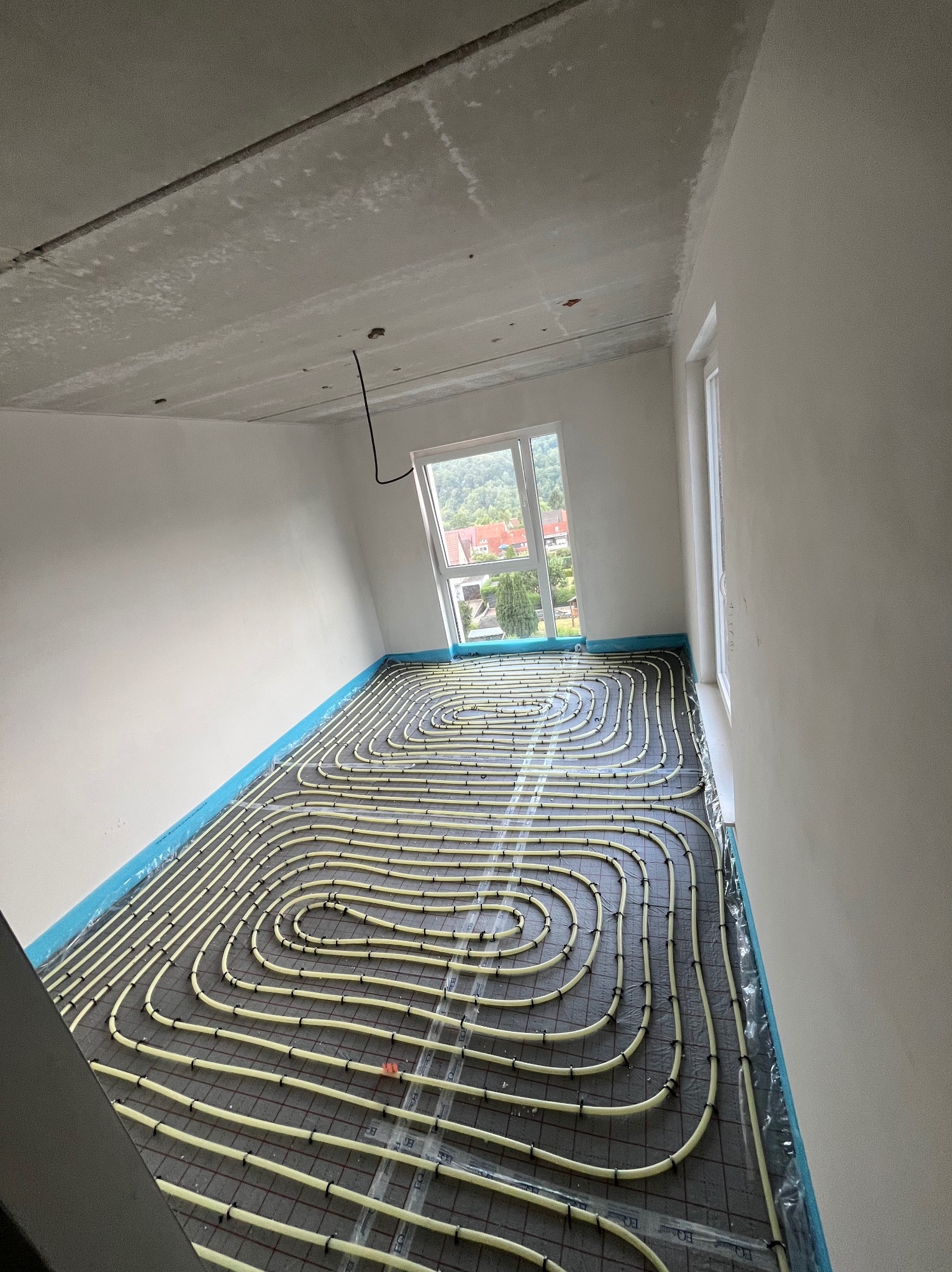 Żółte rury ogrzewania podłogowego ułożone spiralnie na czarnej macie w jasnym pokoju z oknem. Widok na zielone wzgórza przez okno. Niebieska folia na ścianach.