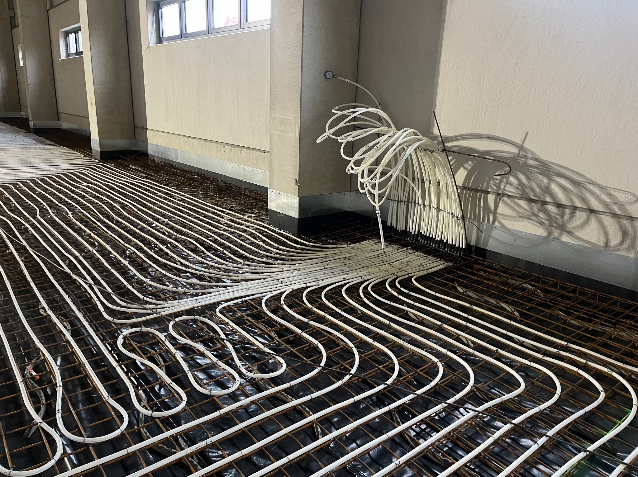 Białe rury ogrzewania podłogowego ułożone na czarnej folii i metalowej siatce w surowym wnętrzu. Widoczne pionowe rury przy ścianie i cień na ścianie.