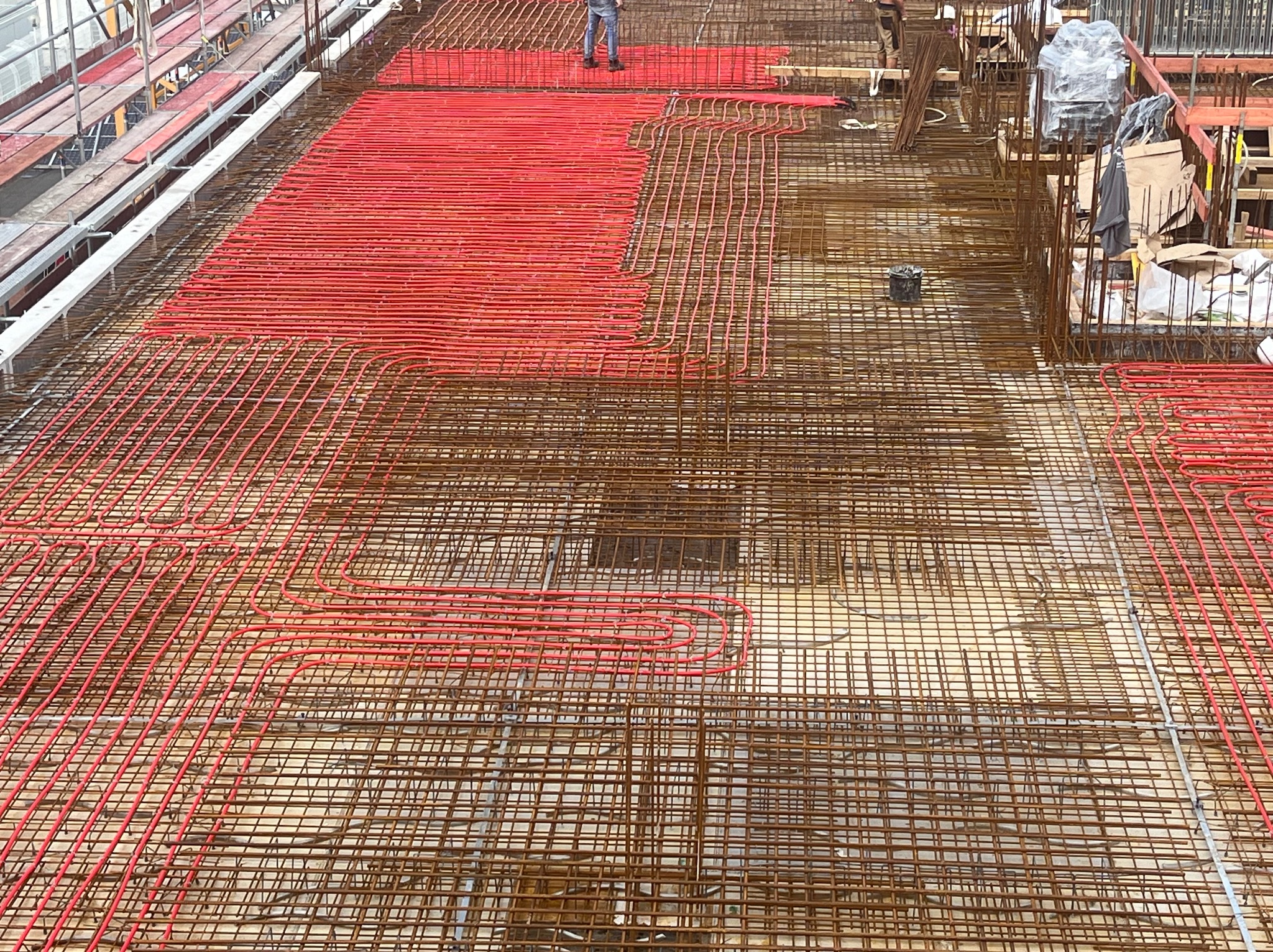Częściowo ułożone, czerwone rury ogrzewania podłogowego na metalowej siatce zbrojeniowej. Widoczne fragmenty rusztowania i osoby w tle.