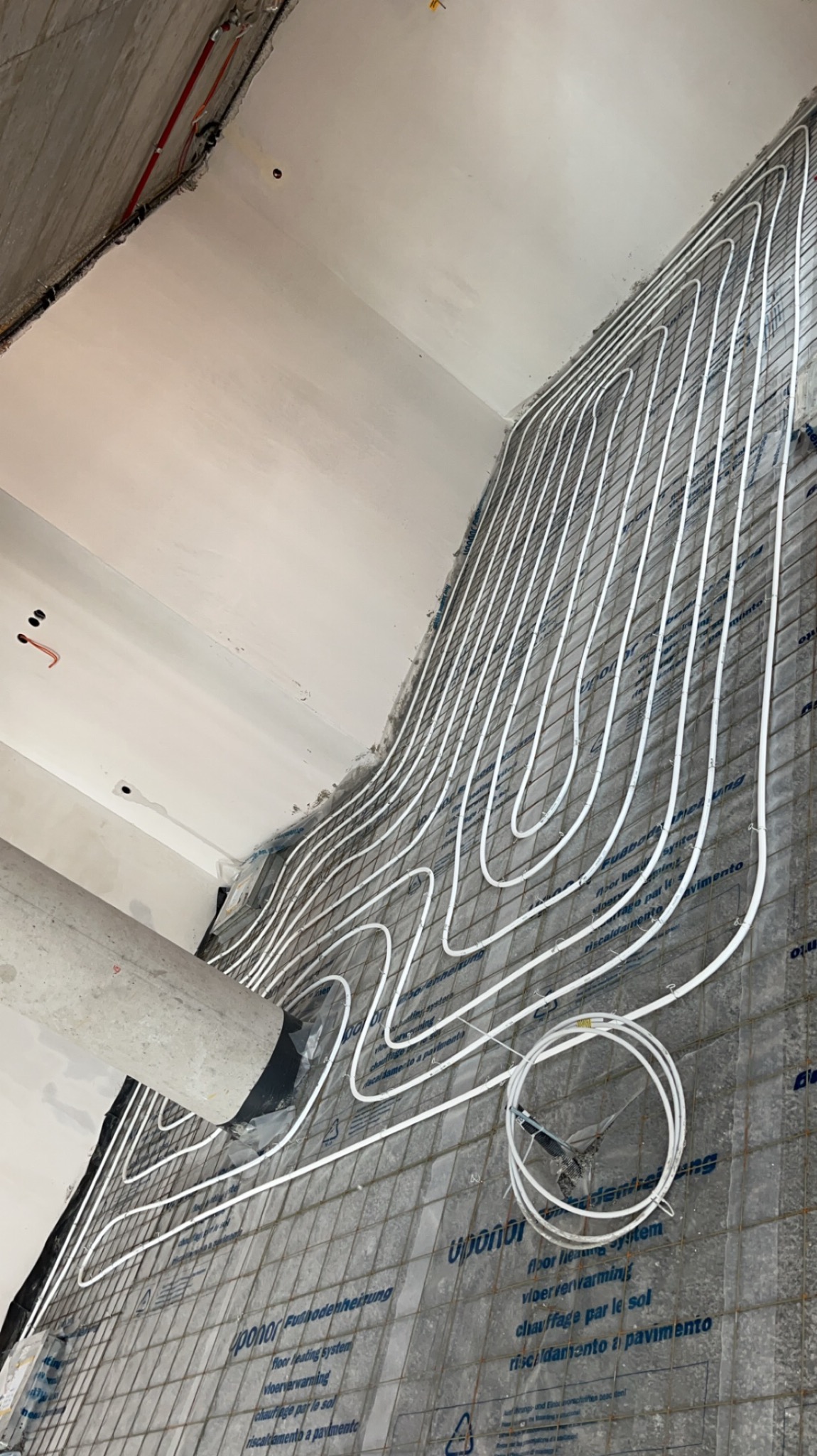 Białe rury ogrzewania podłogowego ułożone na siatce, widoczne elementy instalacji i betonowe ściany w surowym stanie. Ujęcie z dołu, w trakcie montażu.