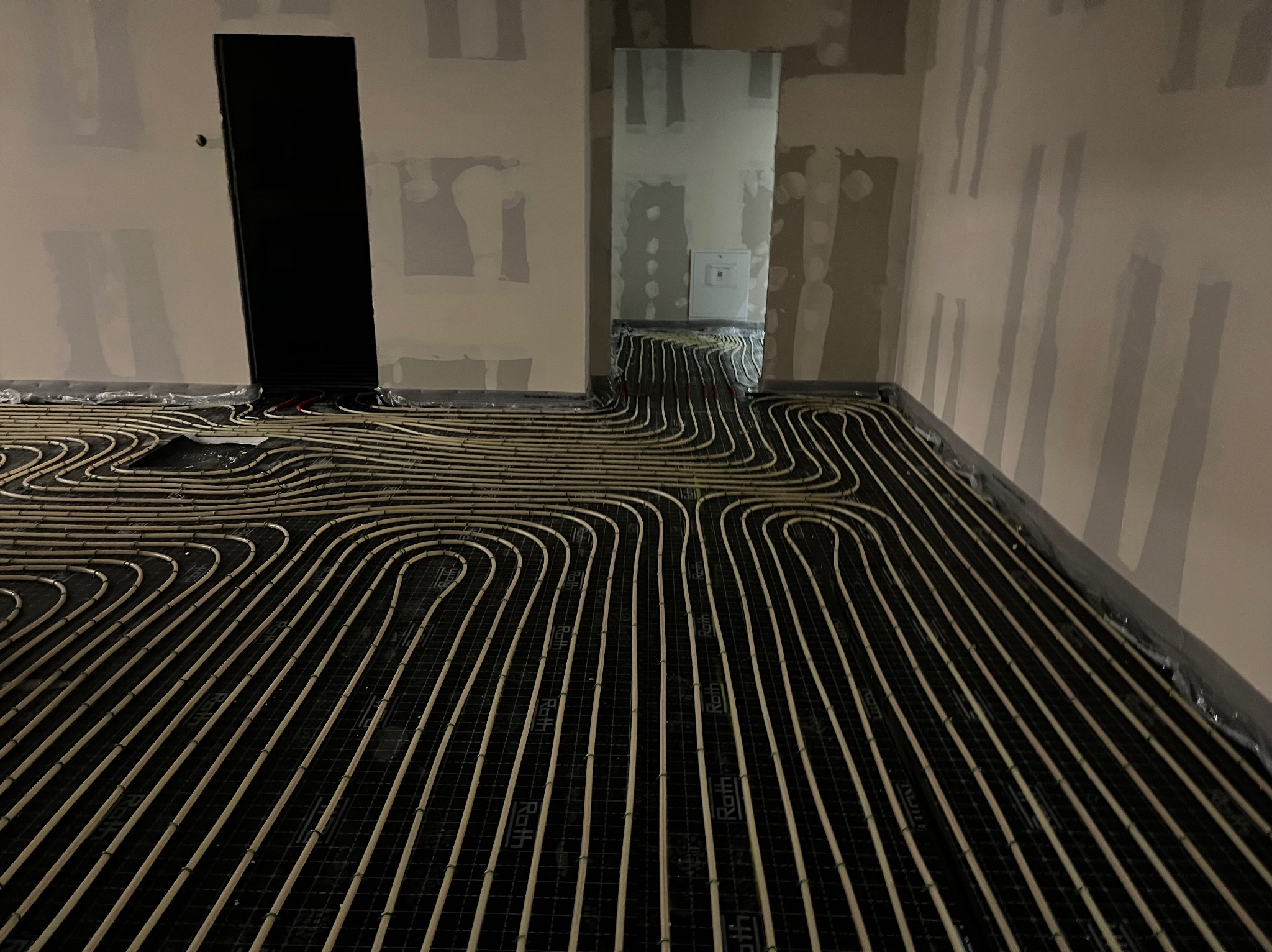 Ułożone rury ogrzewania podłogowego Roth na czarnej macie w surowym wnętrzu. Widoczne ślady po gładzi na ścianach i instalacja pieca grzewczego w tle.
