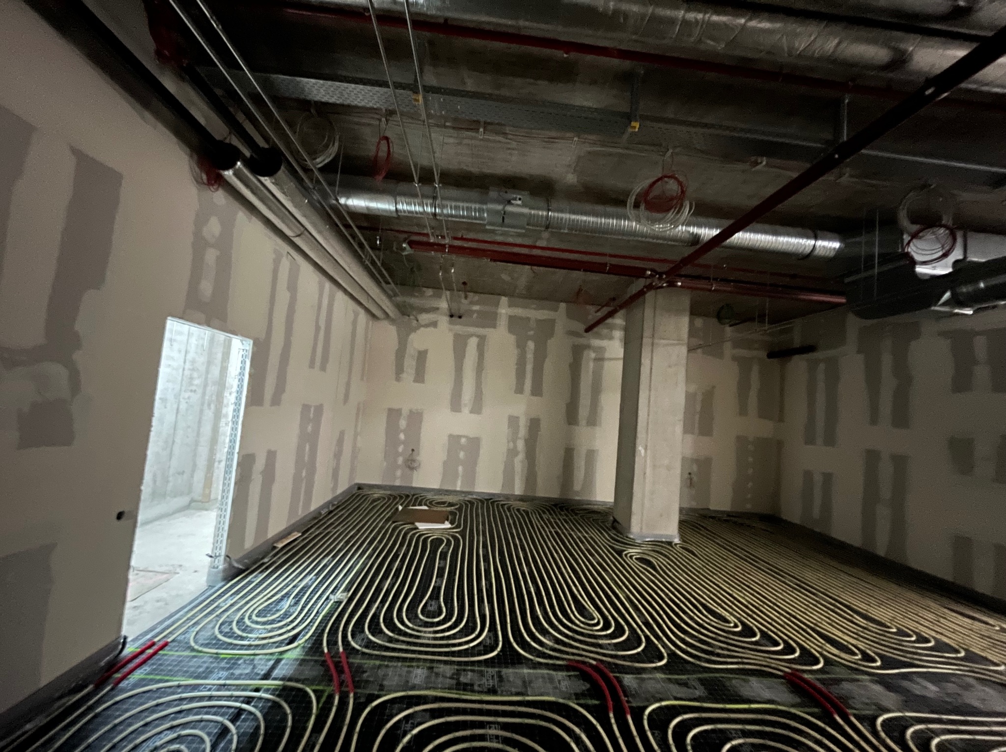 Instalacja ogrzewania podłogowego w surowym wnętrzu. Rury ułożone na czarnej macie, ściany w stanie deweloperskim, widoczne instalacje pod sufitem.