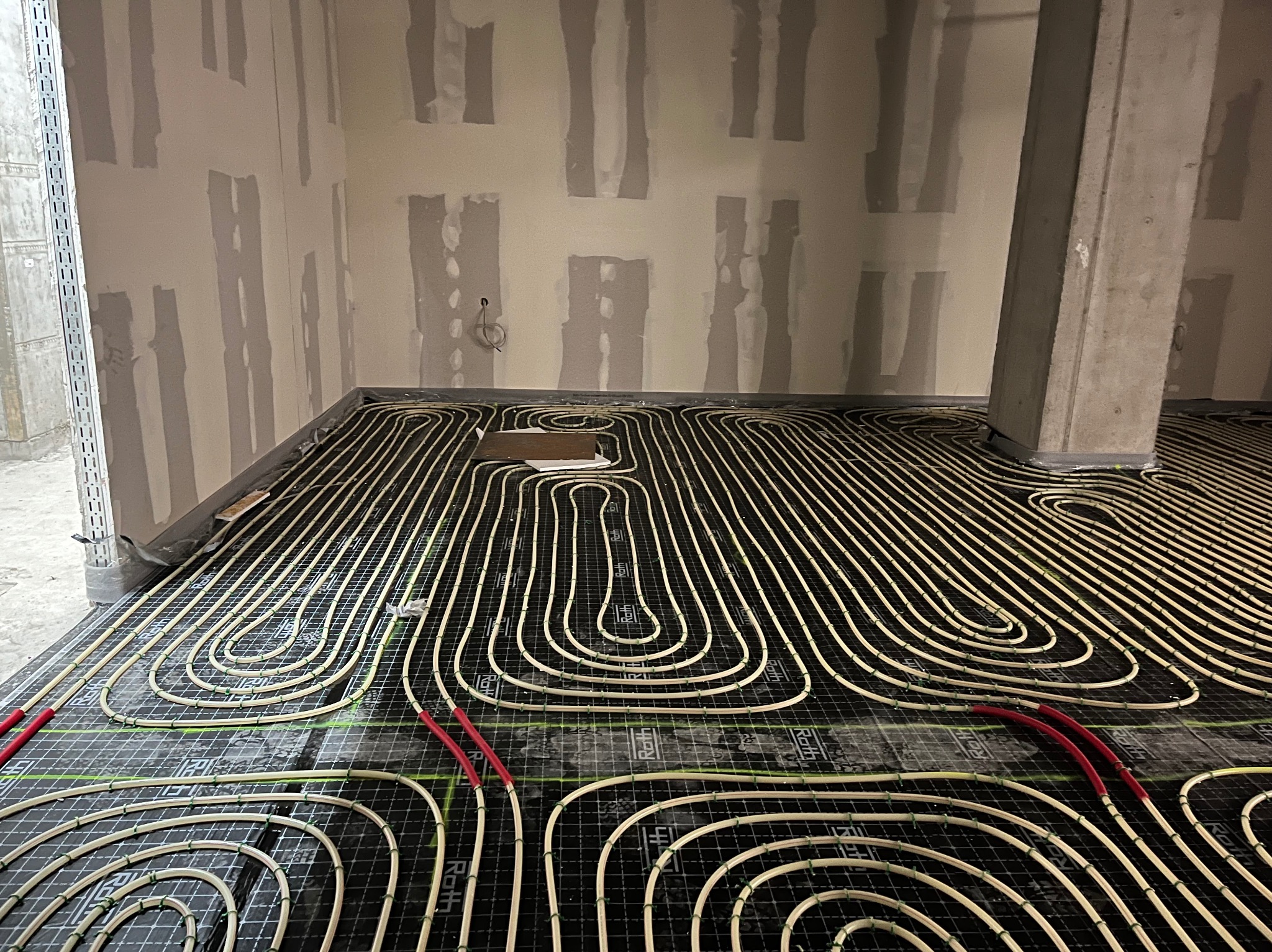 Ułożone rury ogrzewania podłogowego na czarnej macie w surowym wnętrzu z widocznym betonowym słupem i przygotowanymi ścianami do wykończenia.
