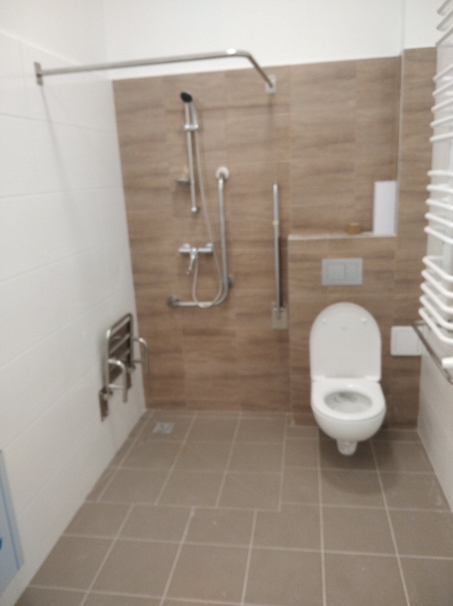 Wykończona łazienka z brązowymi płytkami, toaletą, prysznicem z uchwytem dla niepełnosprawnych i grzejnikiem drabinkowym. Widok na całe pomieszczenie.