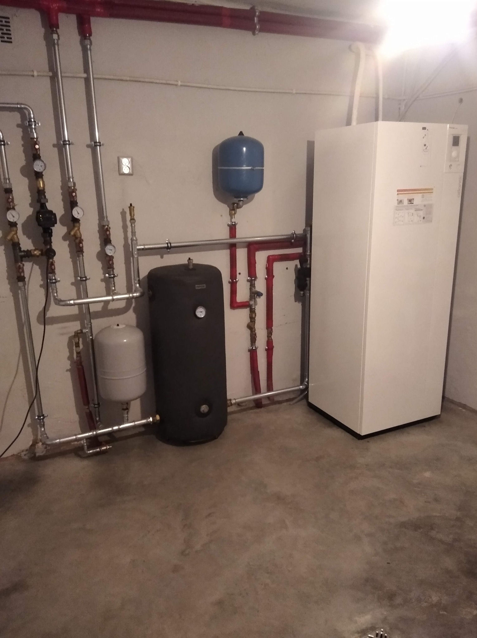 Nowoczesna kotłownia z białym piecem, zbiornikami wyrównawczymi (niebieski i szary) oraz czarnym zasobnikiem ciepłej wody. Widoczne rury miedziane i stalowe.