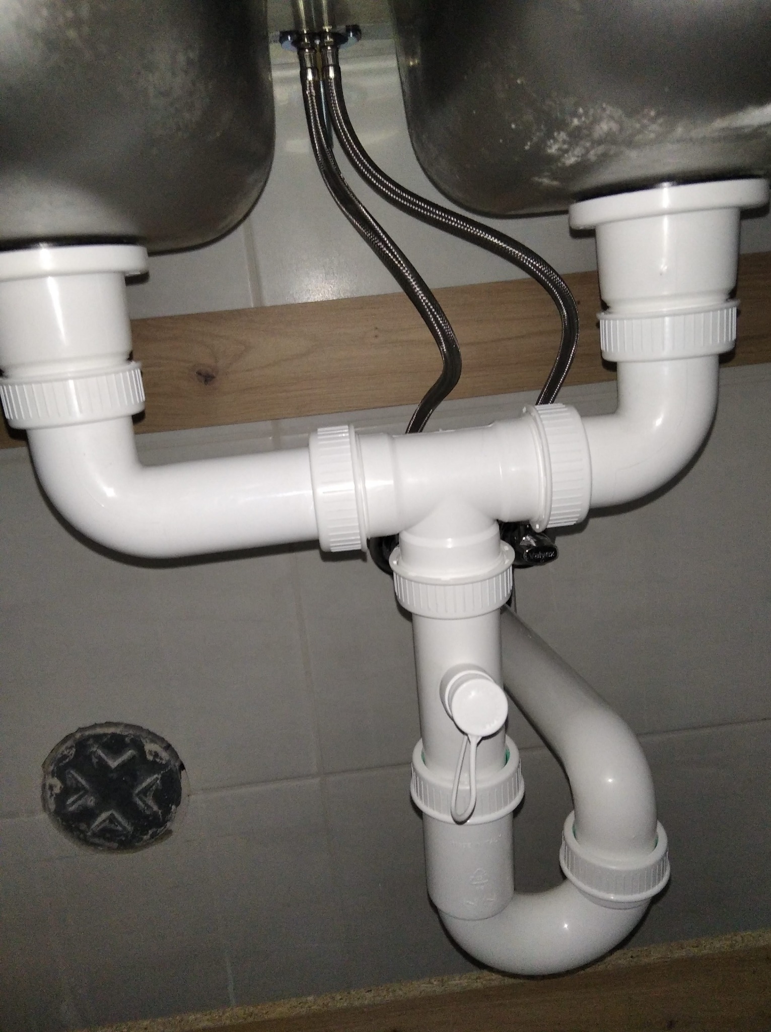 Biały syfon podwójnego zlewu z metalowymi wężykami, widok z dołu, na tle płytek. Uwagę zwraca czarny element w podłodze po lewej stronie.