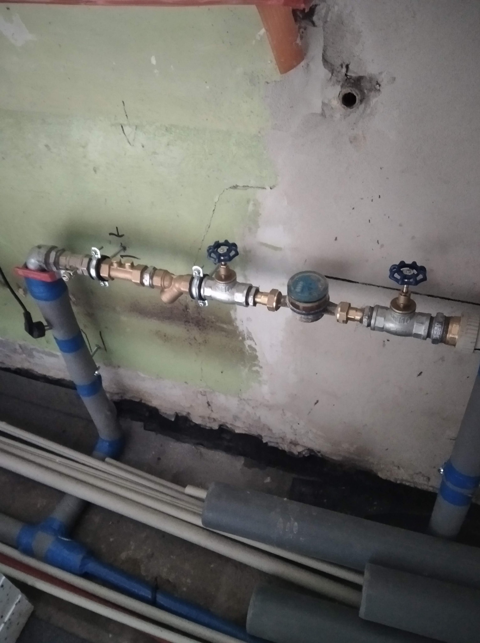 Szczegółowe ujęcie instalacji wodnej z licznikiem, zaworami i rurami, zamontowanej przy ścianie z widocznymi śladami po pracach remontowych.