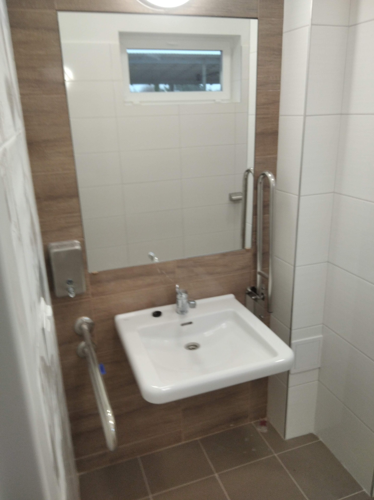 Biała umywalka z chromowaną baterią i uchwytami dla niepełnosprawnych w łazience wykończonej brązowymi i białymi płytkami. Widoczne lustro i dozownik mydła.