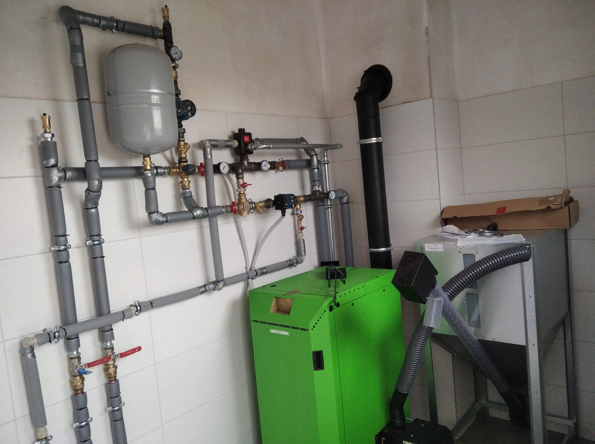 Zielony piec Kostrzewa z szarymi rurami instalacji grzewczej w kotłowni z białymi płytkami. Widoczne zawory, manometry i zbiornik wyrównawczy.