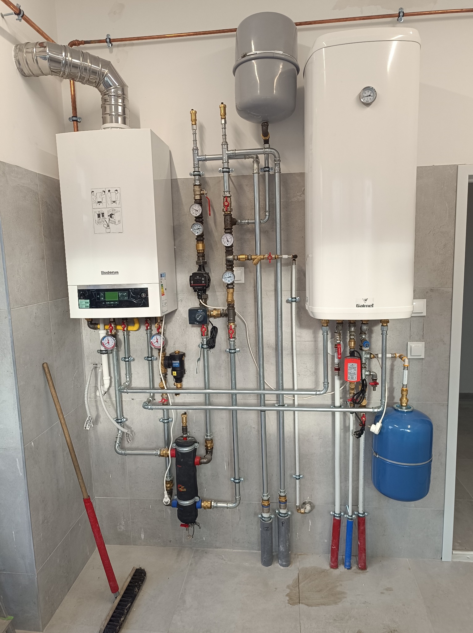 Nowoczesna kotłownia z białym piecem gazowym Buderus i zasobnikiem Galmet, rurami, manometrami. Widoczny filtr i naczynie przeponowe. Ściany i podłoga wyłożone szarą płytką.